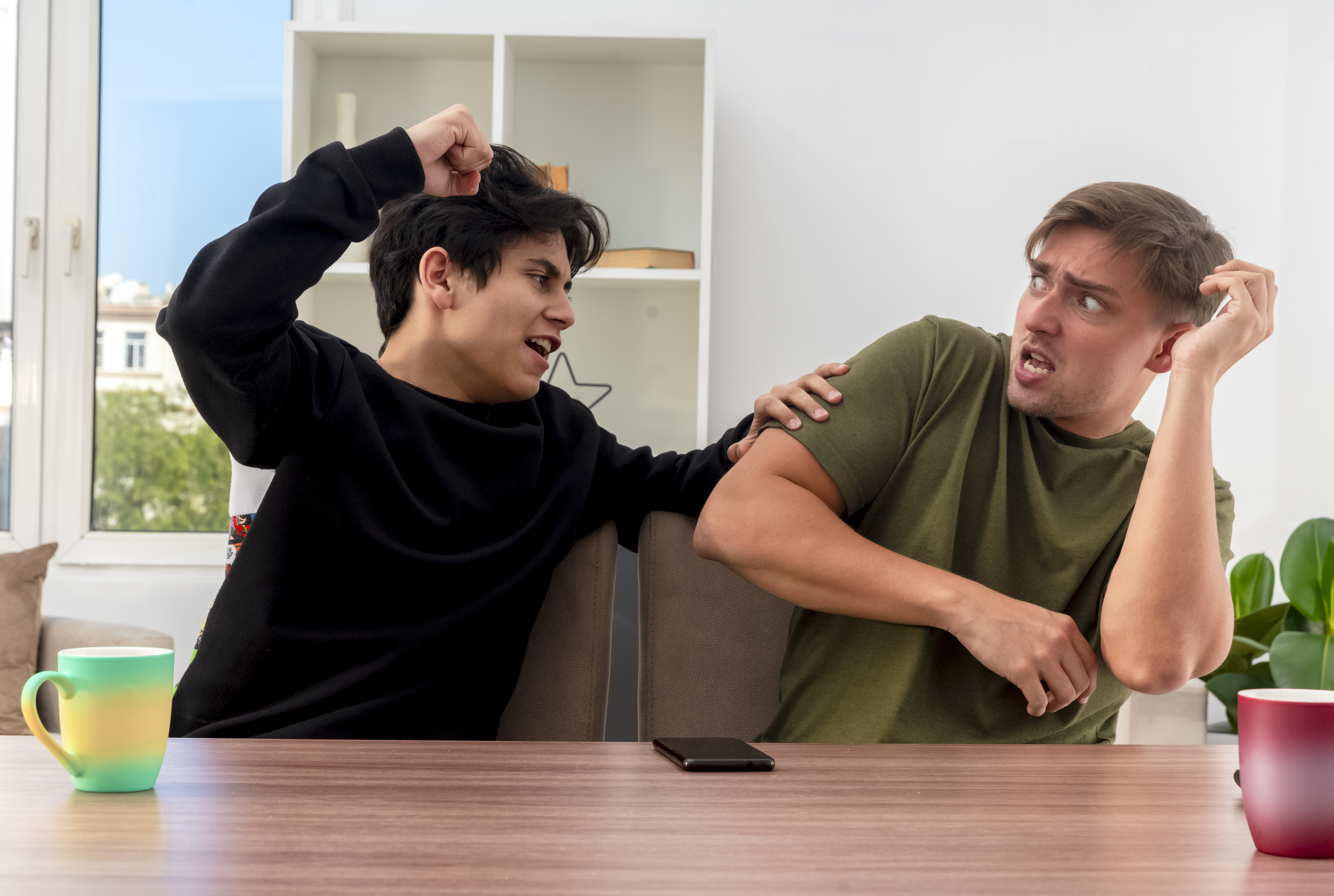 scared-young-blonde-handsome-man-raising-hand-sitting-table-looking-furious-young-brunette-handsome-boy-hitting-him.jpg